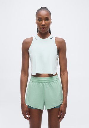 Αθλητική γυναίκα με αμάνικο cropped τοπ σε ανοιχτό μέντα και αντίστοιχα σορτς για τρέξιμο, στέκεται μπροστά σε λευκό φόντο.