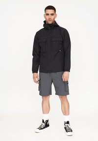 Schwarze wasserdichte Jacke mit zwei Brusttaschen, kombiniert mit grauen Cargo-Shorts. Modell trägt schwarze High-Top-Sneaker mit weißen Sohlen.