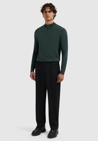Donkergroene lange mouw top met een halve rits kraag, gecombineerd met losse zwarte broek en zwarte schoenen. Eenvoudig, glad ontwerp met minimale details.