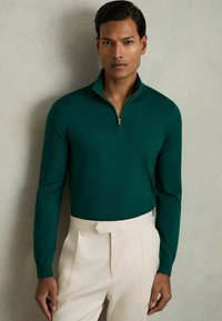 Pull-over vert zippé en tissu doux avec des manches longues, associé à un pantalon crème doté d'une taille plate et de plis.