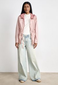Chaqueta rosa claro con capucha y cordones, combinada con unos vaqueros de mezclilla sueltos y azul claro. El atuendo presenta líneas simples y limpias, y un ajuste relajado.