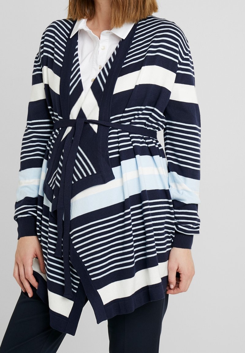 Femme portant un cardigan rayé bleu marine, blanc et bleu clair avec ceinture, sur une chemise blanche à col et un pantalon noir, debout devant un fond gris.