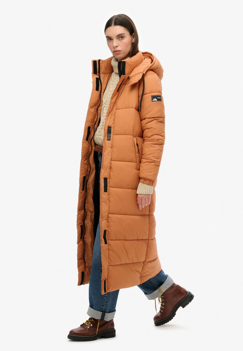 Lange oranje pufferjas met zwarte accenten en capuchon, voorzien van een gestikt ontwerp en voorzakken, gedragen over een crèmekleurige trui en blauwe jeans.