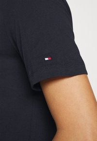 Černé bavlněné tričko s těsným střihem. Na rukávu se nachází malé vyšívané logo v červené, bílé a modré barvě. Hladká textilie.