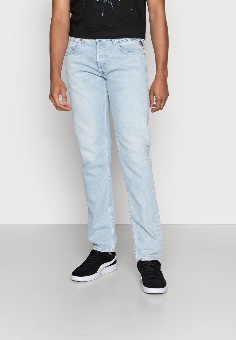 Ljusblå denimjeans med rak passform, som har fem fickor och diskret sömnad, ihop med svarta sneakers på en ljusgrå bakgrund.