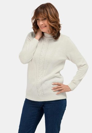 Frau mit schulterlangem braunem Haar, die lächelt und einen cremefarbenen Rollkragenpullover aus Strick sowie dunkelblauen Jeans trägt, posiert mit der Hand auf der Hüfte.