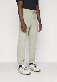 Pantalones verde oliva claros con cintura elástica y cordón, que cuentan con bolsillos laterales y puños ajustados, fabricados en tejido ligero.