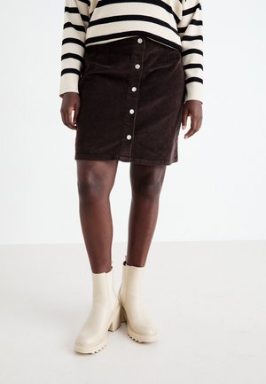 Personne portant une jupe en velours côtelé marron foncé à boutons sur le devant, des bottines épaisses crème et un pull crème à rayures horizontales noires.