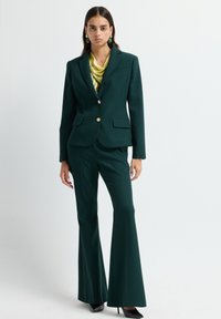 Groene op maat gemaakte suit met een gestructureerde blazer, gouden knopen en flared broekspireeën. Bevat een zijden gele sjaal en zwarte hoge hakken.