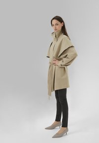 Bézs öves ballonkabát magas gallérral és túlnagyított ujjakkal, fekete karcsúsított nadrággal és világosszürke hegyes orrú cipővel párosítva.