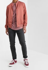 Chaqueta bomber rosa claro con botones a presión, llevada sobre una camisa a juego, combinada con jeans oscuros y zapatillas burdeos.
