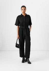 Mono de lino negro con mangas cortas, cinturón en la cintura y bolsillos en el pecho. Combinado con botas negras de tacón ancho y un pequeño bolso negro.