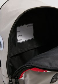 De binnenkant van de grijze rugzak heeft een zwarte voering, een naamlabel, opbergvakjes en een zichtbare ritssluiting met rode accenten.