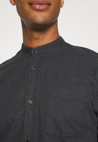 Hombre vistiendo una camisa de color gris oscuro con cuello mandarín, botones marrones y un bolsillo en el pecho, mostrado desde la barbilla hasta la parte superior del torso.