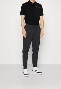 Svart pikétröja med broderad "Polo"-logotyp, tillsammans med mörkgrå sweatpants med justerbar dragsko, och vita sneakers med svarta accenter.