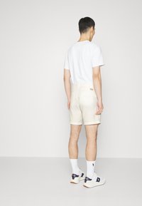 Svetlé béžové šortky s hladkou texturou, ohrnutými manžetami a detailem štítku na zadní straně, doplněné bílým tričkem a bílými ponožkami.