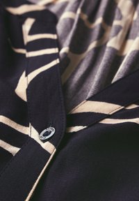 Camicia con bottoni caratterizzata da un motivo a righe nere e beige, realizzata in tessuto leggero con una texture liscia. È visibile un'accortezza in metallo sul bottone.