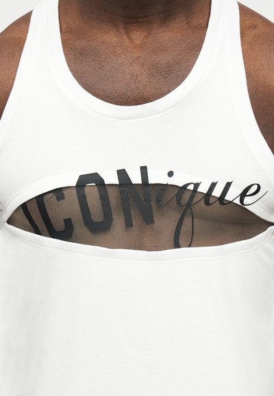 Κοντινό πλάνο του άνω μέρους του στήθους ενός ατόμου που φοράει λευκό μπλουζάκι χωρίς μανίκια με οριζόντια εγκοπή που αποκαλύπτει μαύρο δίχτυ και εν μέρει ορατό μαύρο κείμενο από κάτω.