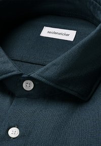 Camicia in cotone verde scuro con colletto classico, caratterizzata da due bottoni bianchi e un'etichetta del marchio cucita nel colletto. Tessuto morbido.
