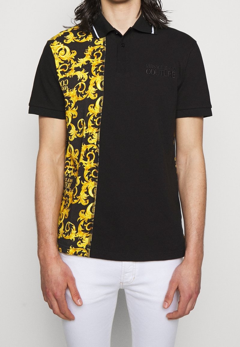 Homme portant un polo noir à manches courtes avec un imprimé baroque doré sur le côté gauche, associé à un pantalon blanc, debout devant un fond blanc.