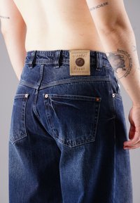 Person wearing dark blue jeans with a leather label reading "Picaldi Jeans Authentic Design," showing tattoos on arms and a bare back.