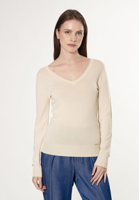 Cremfarbener V-Ausschnitt-Pullover mit langen Ärmeln in taillierter Passform, aus glattem Material mit gerippten Bündchen, kombiniert mit blauen strukturierten Hosen.