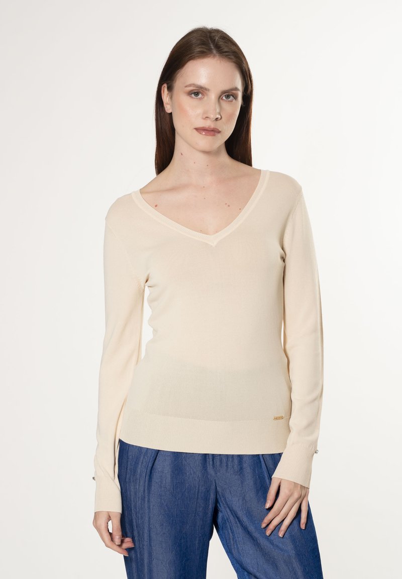 Cremfarbener V-Ausschnitt-Pullover mit langen Ärmeln in taillierter Passform, aus glattem Material mit gerippten Bündchen, kombiniert mit blauen strukturierten Hosen.