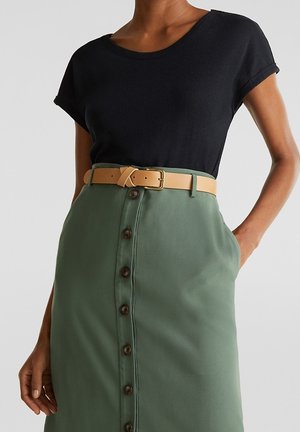 Kvinde iført en sort kortærmet top, der er stoppet ned i et grønt nederdel med knapper foran og et beige bælte, med hænderne afslappet ved siderne.