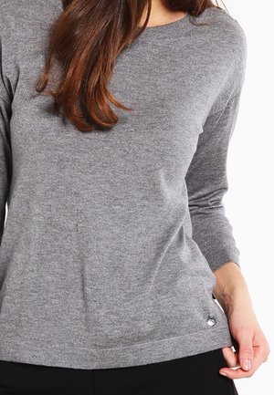Femme portant un pull gris ajusté à manches longues avec un col rond, touchant l’ourlet sur son côté droit.