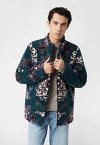 Teal spalvos fliso striukė su baltais ir bordo gėlių raštais, aukštu apykakle, visiškai užtraukiama priekyje ir dviem kišenėmis. Minkšta tekstūra.