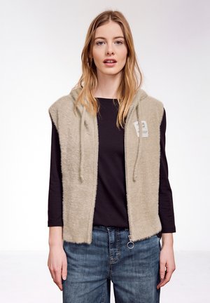 Jeune femme aux longs cheveux blonds portant un gilet beige à capuche zippé sans manches par-dessus un t-shirt noir à manches longues et un jean bleu, debout droite.