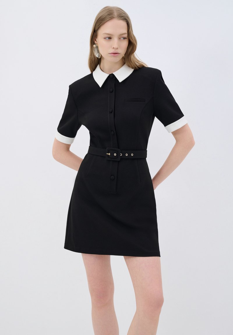 Schwarzes Kleid mit kurzen Ärmeln und weißem Kragen, mit tailliertem Schnitt, schwarzem Gürtel und Knopfverschluss, aus strukturiertem Stoff gefertigt.