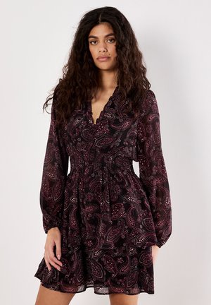 Frau mit langen lockigen Haaren, die ein schwarzes Kleid mit pinkem Paisley-Muster, langen Ärmeln und einem taillierten Bund trägt, steht vor einem schlichten Hintergrund.