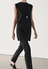 Personne marchant à l'intérieur portant une tunique noire sans manches, un pantalon noir, des chaussures noires et un collier pendentif devant un mur blanc uni.