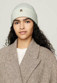 Berretto lavorato a maglia grigio chiaro con texture a coste e un piccolo logo in metallo, abbinato a un cappotto marrone a motivo spina di pesce e un maglione color crema.