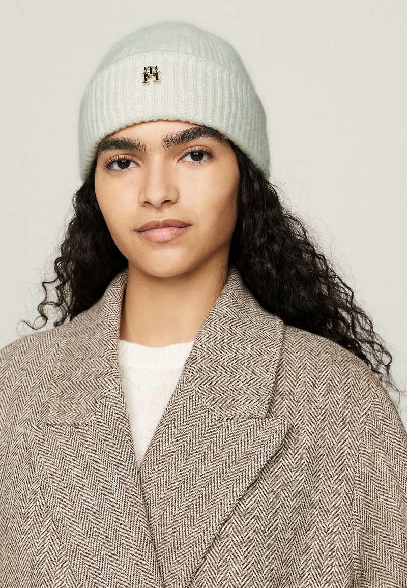 Berretto lavorato a maglia grigio chiaro con texture a coste e un piccolo logo in metallo, abbinato a un cappotto marrone a motivo spina di pesce e un maglione color crema.