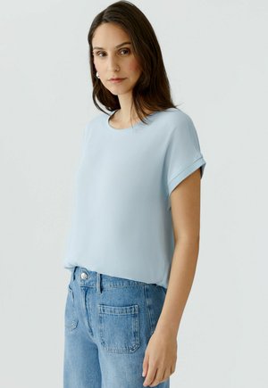 Frau trägt ein hellblaues kurzärmeliges Oberteil und hoch taillierte blaue Jeans, steht vor einem schlichten weißen Hintergrund.