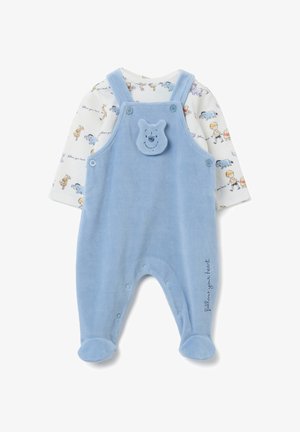 Romper de bebé azul claro con pies y tirantes a botones, con una cara de oso sonriente en el pecho, superpuesto a una camiseta blanca de manga larga con personajes de dibujos animados.