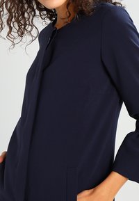 Žena v dobře padnoucím tmavě modrém kabátu s dlouhým rukávem a skrytým zavíráním vpředu, s rukou v kapse a částí kudrnatých vlasů viditelných.