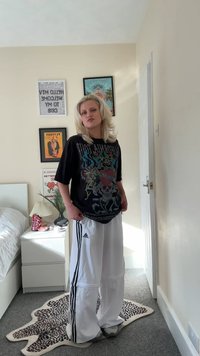 Svart grafisk t-skjorte med skallemotiv, kombinert med hvite Adidas trackbukser med svarte striper. Mot en nøytral rombakgrunn.