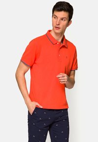 Męska koszulka polo, jasnopomarańczowa z kontrastowym niebieskim kołnierzykiem, krótki rękaw i dopasowany fason. W połączeniu z granatowymi spodniami z wzorem rowerów.