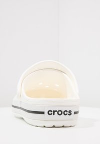 Hvite Crocs tøysko med buet bakdel, åpen tå, glatt plastoverflate og svart aksentstripe med logo på hælen.