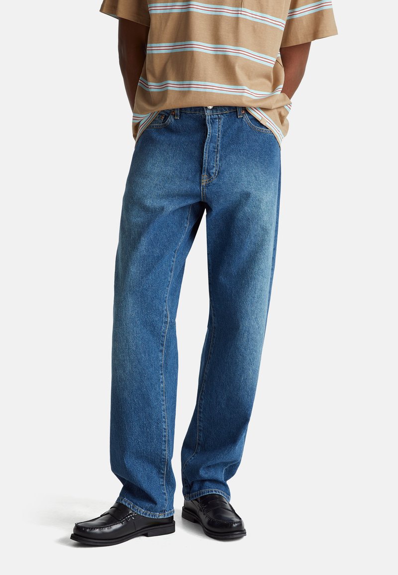 Niebieskie dżinsy o luźnym kroju i prostych nogawkach, z detalicznym przeszyciem i wyblakłą teksturą, w połączeniu z czarnymi butami.