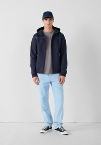 Marine hoodiejack met een gestructureerde schouderpartij, gedragen over een grijze shirt. Lichtblauwe jeans en zwarte sneakers maken de look compleet. De pet past bij de jas.