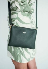 Zelená kožená crossbody taška se zipovým uzávěrem, s drobným zlatým logem a úzkým popruhem, zobrazující elegantní obdélníkový design.