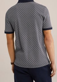 Námořnická modrá polo tričko s geometrickým vzorem a krémovými a rezavými akcenty, krátkými rukávy a pevným námořnickým límcem a manžetami.