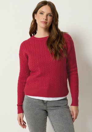 Femme aux longs cheveux bruns portant un pull en maille torsadée rouge par-dessus une chemise blanche et un jean gris, debout devant un fond uni.