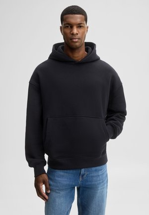 Mann trägt einen schwarzen Kapuzenpullover mit Fronttasche und blaue Jeans, steht vor einem einfarbigen hellgrauen Hintergrund.