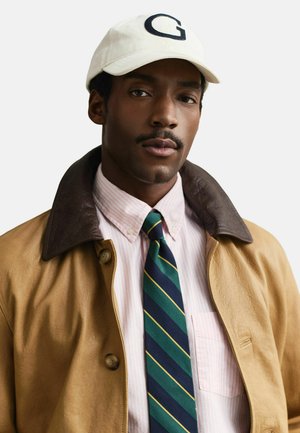 Man met een witte pet met de letter G, een beige jas met een donkere kraag, een roze gestreept overhemd en een groen, marineblauw en geel gestreepte stropdas.