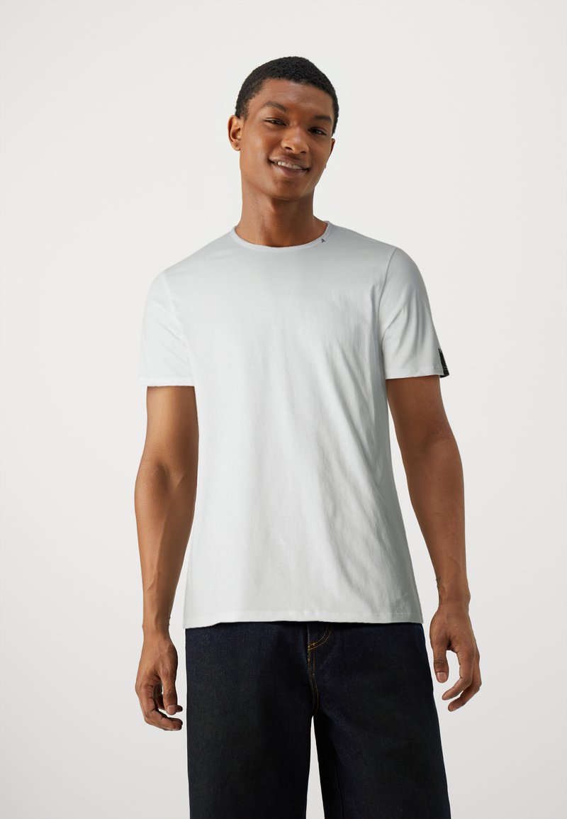 Hvid bomulds T-shirt med korte ærmer, rund halsudskæring og en sort detalje på ærmet, båret med mørkeblå jeans.